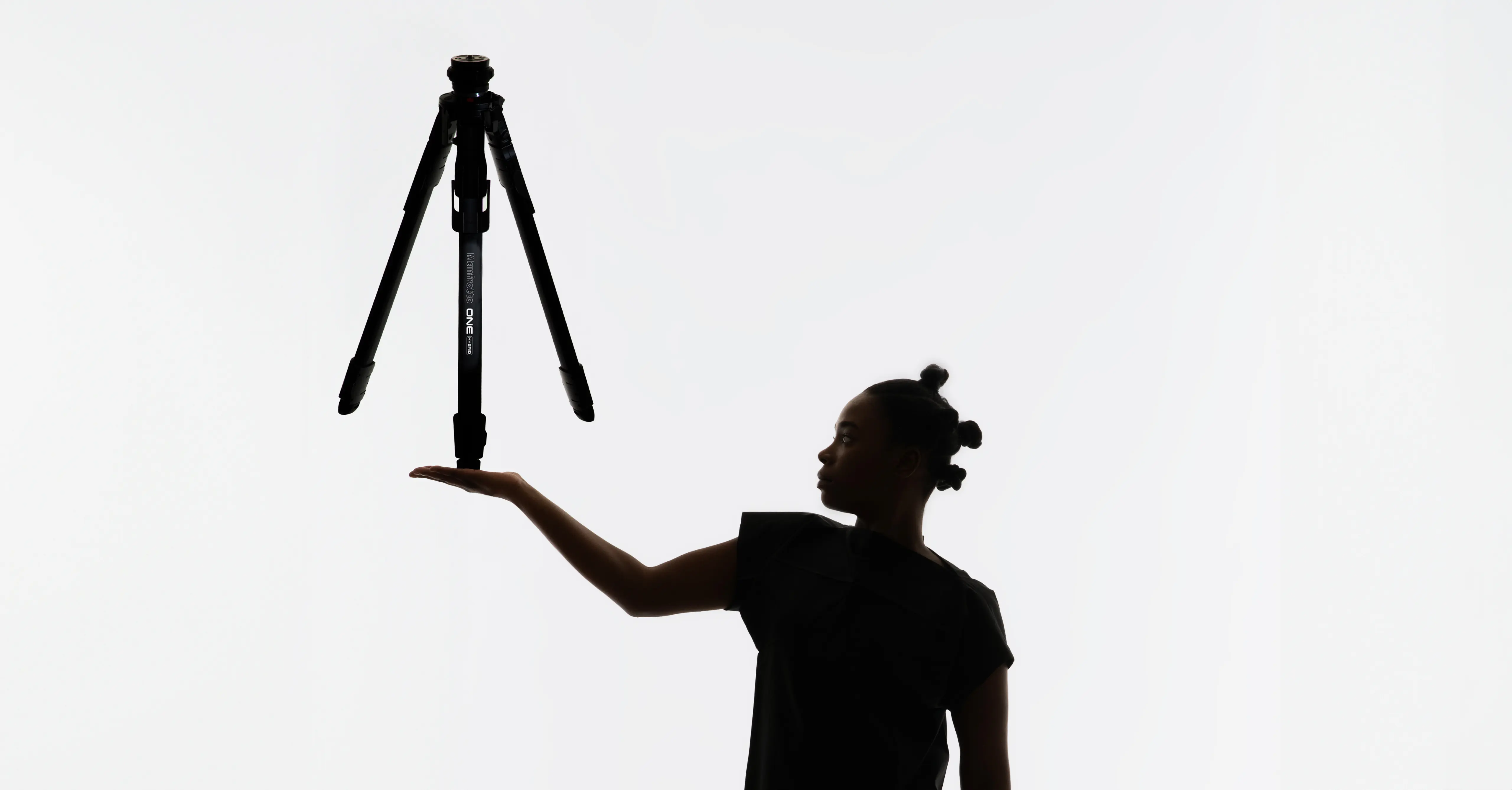 A person stands against a white background, holding the ONE tripod Close-up of the ONE tripod by leading industrial design consultancy LAYER showing the tripod head and built-in spirit level on a white background.balanced above their open hand.